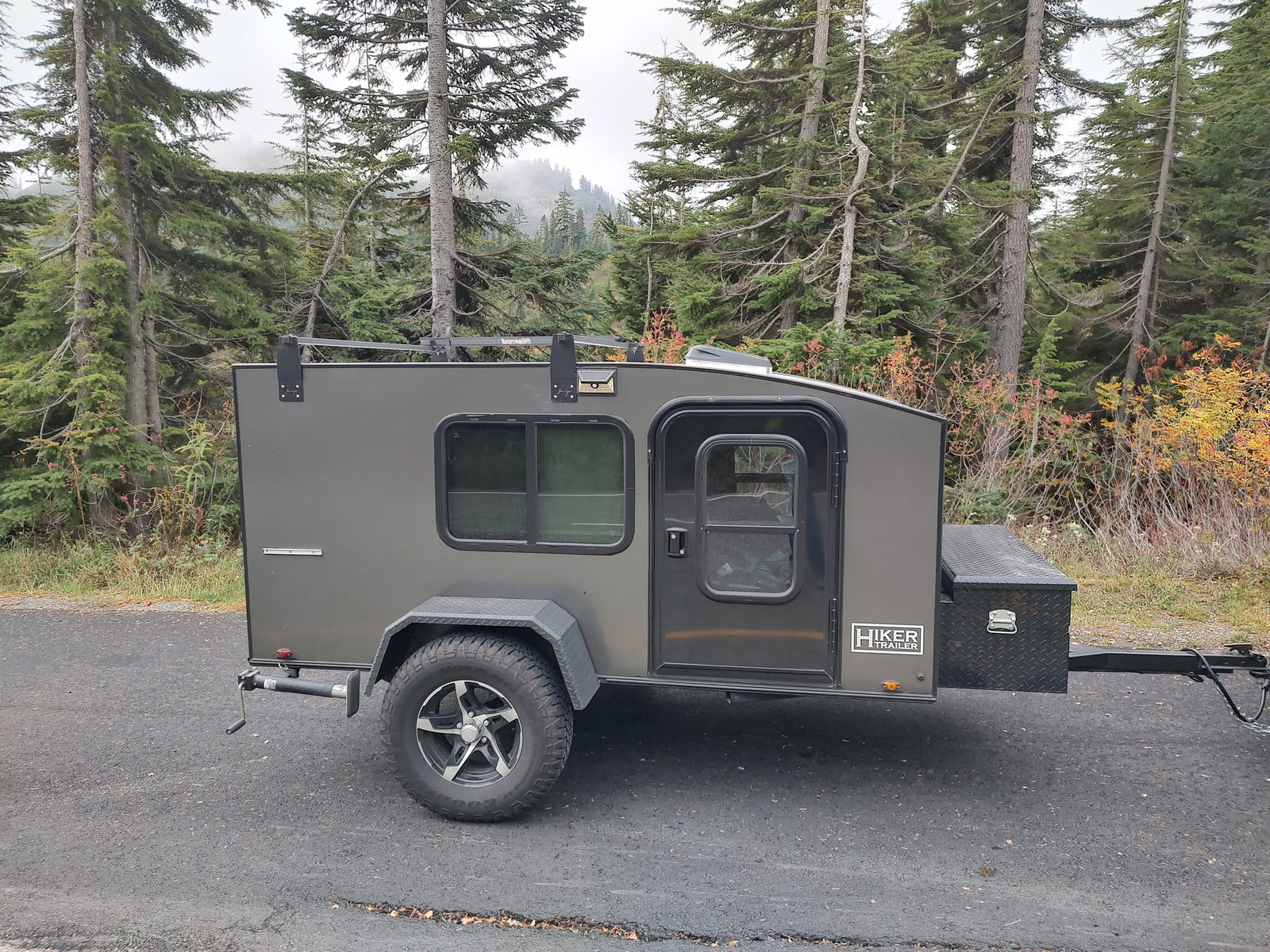 This gray Hiker Trailer RV rental is parked on asphalt, framed by tall evergreen trees and autumn leaves.