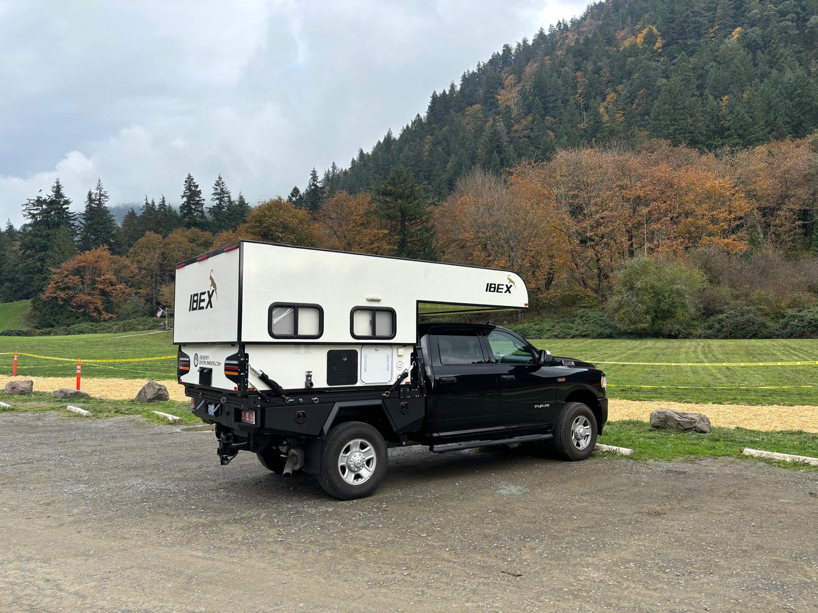 Black Ram truck with white IBEX truck camper parked by mountains. Ideal RV rental for exploring rugged landscapes.|Compact RV rental wet bath with wall-mount toilet & shower.|This RV rental offers a comfortable sleeping space with a wide bed