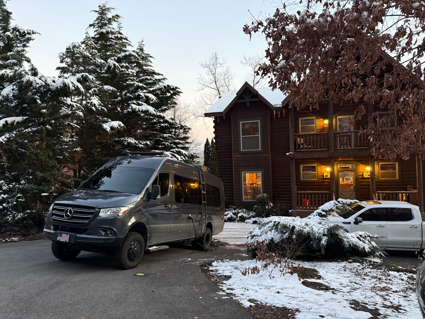 Experience winter! This RV rental, a Mercedes camper van, is parked at a snowy log cabin with evergreens.