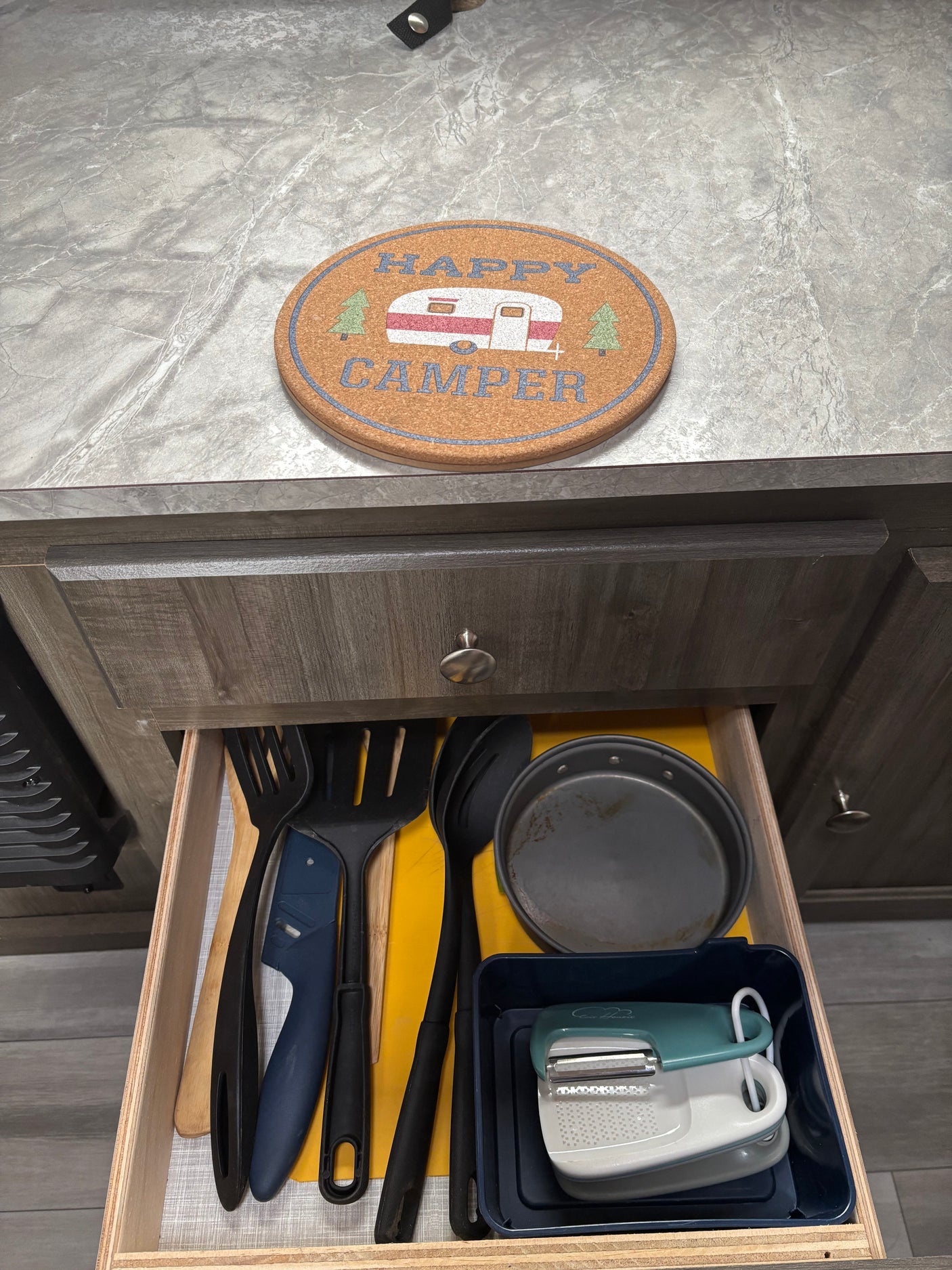 This RV rental's organized kitchen drawer holds cooking utensils, a pan, peeler, and grater, with a "Happy Camper" coaster above.