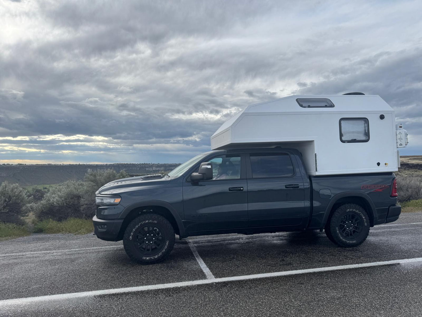 parked on dirt in a rugged landscape under a cloudy sky.|Dark gray Ram pickup with white truck camper. Perfect RV rental for overland travel and camping adventures under cloudy skies.|RV rental truck camper in a grassy spot by a wooden fence