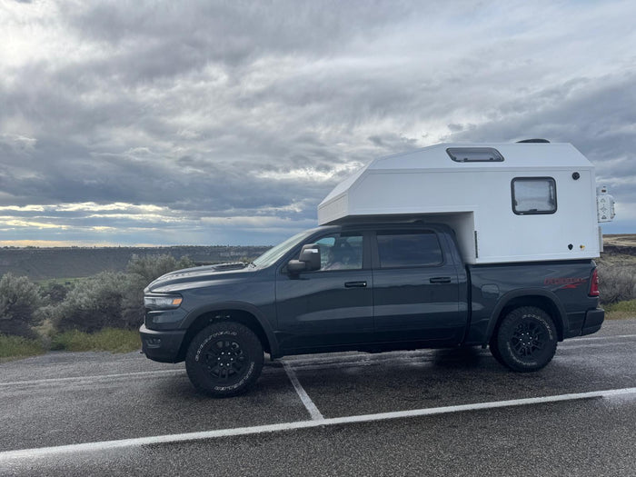 parked on dirt in a rugged landscape under a cloudy sky.|Dark gray Ram pickup with white truck camper. Perfect RV rental for overland travel and camping adventures under cloudy skies.|RV rental truck camper in a grassy spot by a wooden fence
