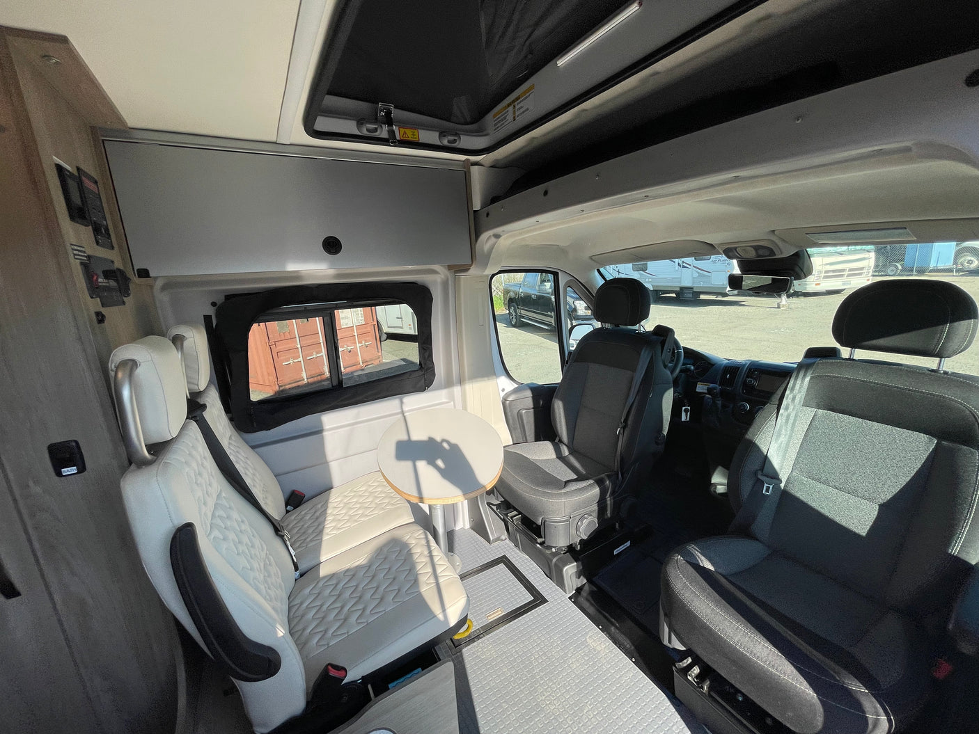 Bright RV rental interior featuring a dinette with cream seats and a table, plus front cabin seats and overhead storage.