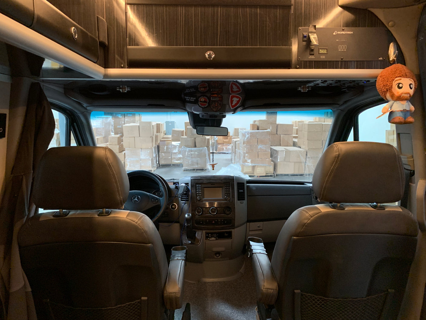 Inside an RV rental: brown front seats and dashboard facing stacks of boxes in a warehouse. A Bob Ross doll hangs.