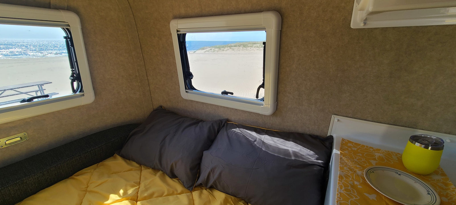 RV rental interior: cozy bed, gray pillows, yellow bedding, and two windows offering serene views of the beach, ocean, and sand dunes.