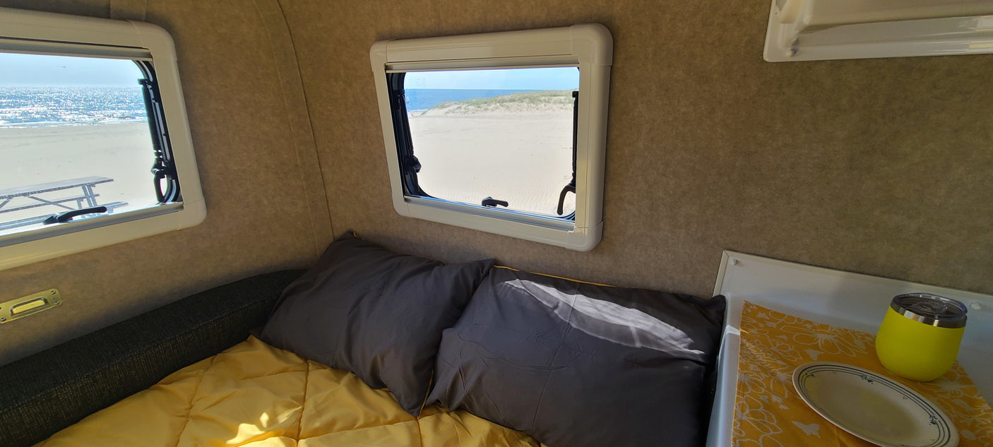 RV rental interior: cozy bed, gray pillows, yellow bedding, and two windows offering serene views of the beach, ocean, and sand dunes.