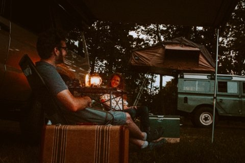 signaling a relaxing outdoor evening.|Discover RV rental adventures! Couple relaxing by a classic 4x4 with a rooftop tent and lantern for evening camping.|Rear of a classic green Land Rover with a black CVT roof tent packed. Camping gear inside
