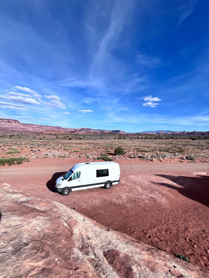 A white RV rental is parked in a vast red desert landscape with distant mesas under a bright blue sky.