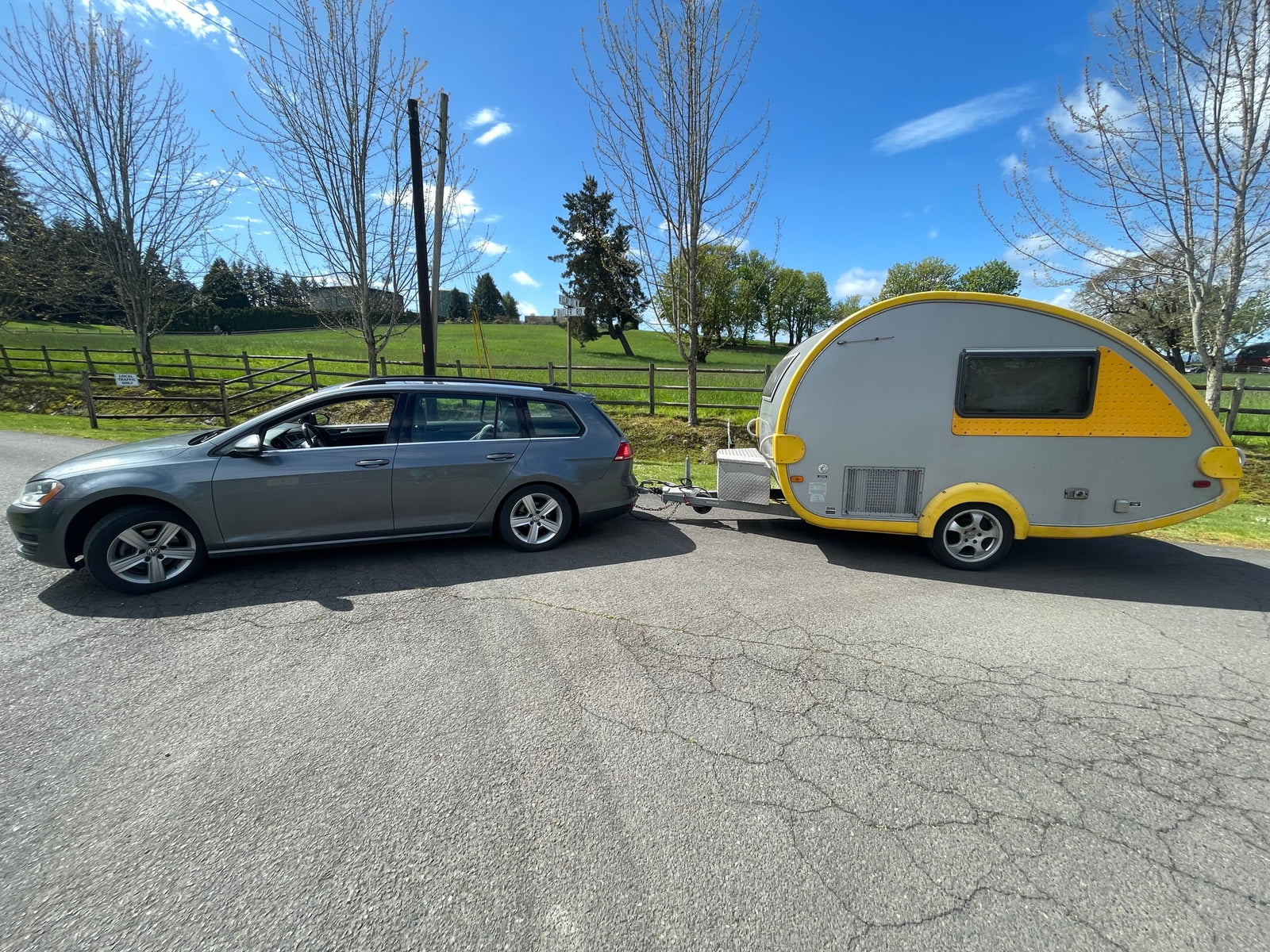 Grey VW station wagon towing a yellow/grey teardrop camper on an asphalt road. Perfect RV rental for your next adventure.