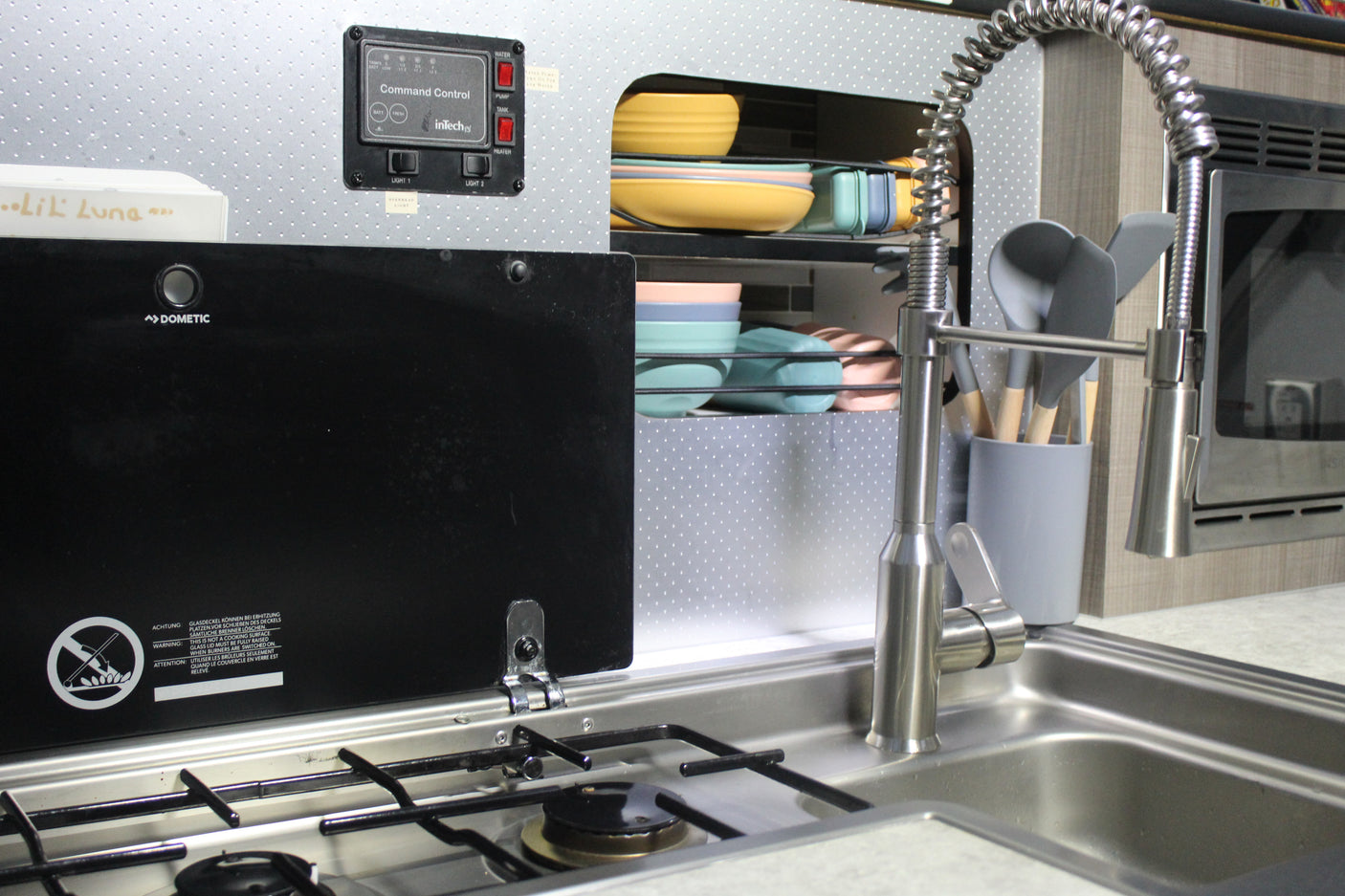 Compact RV rental kitchen with Dometic stove, sink, colorful dishes, and an InTech control panel for water and power.