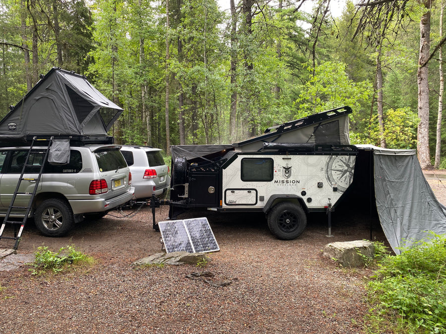 Explore the wilderness! RV rental featuring an SUV with a rooftop tent, overland trailer, and solar power setup in a lush forest.