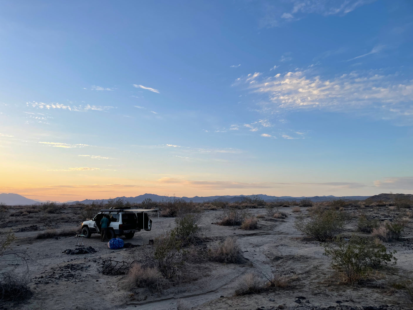 A person prepares camp with an RV rental SUV in the desert at sunset, surrounded by sparse brush & distant mountains.
