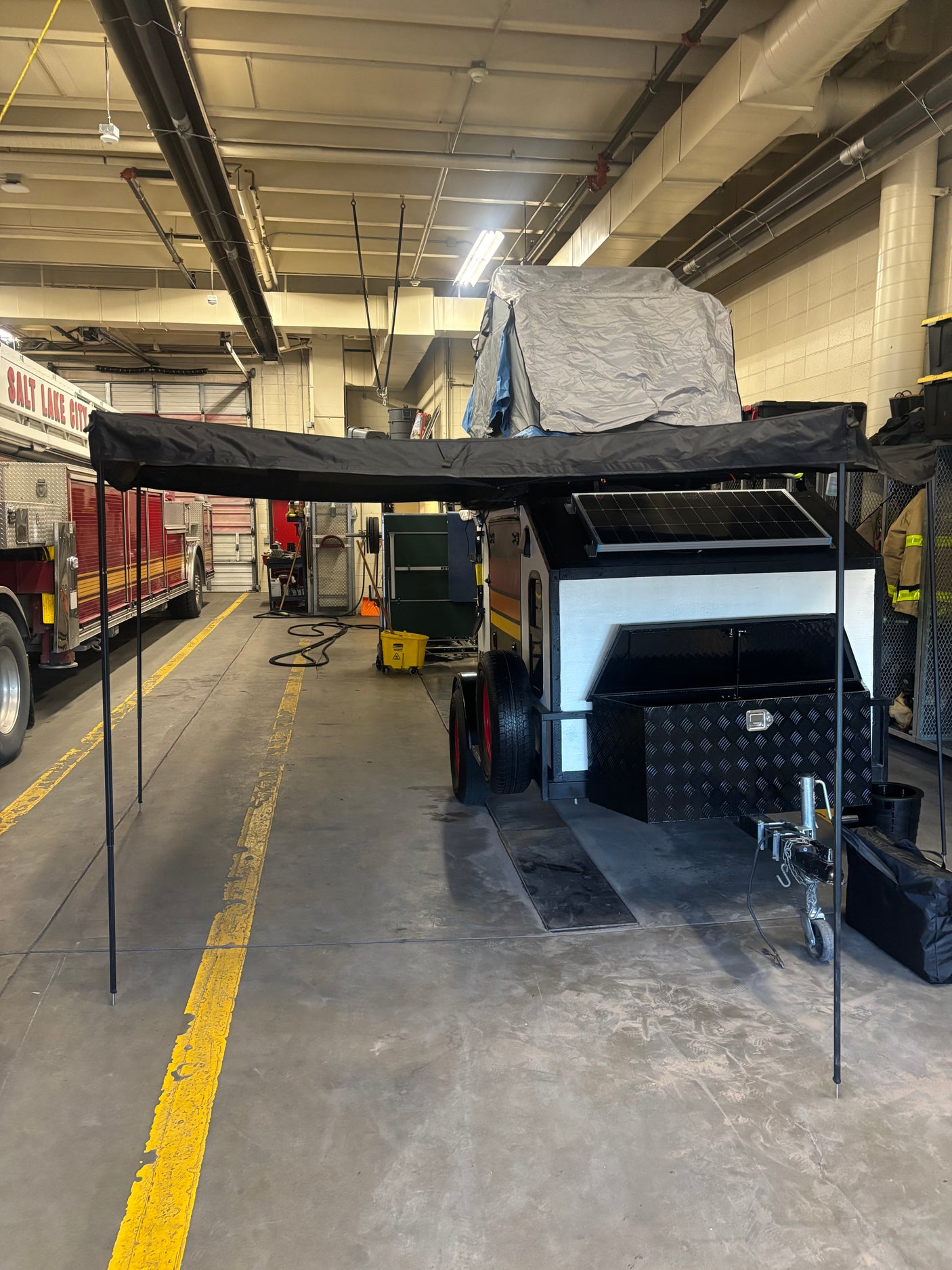 Off-road camping trailer for rv rental, featuring an extended awning, rooftop tent, and solar panels, parked in a large garage.