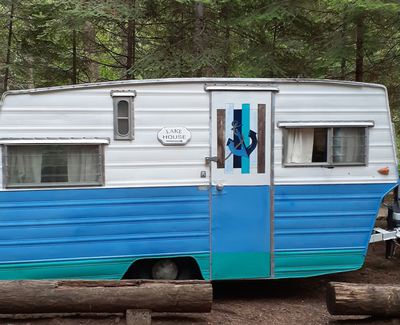 Retro rv rental: This charming blue/teal & white vintage travel trailer features an anchor design & 'LAKE HOUSE' sign, nestled in trees.