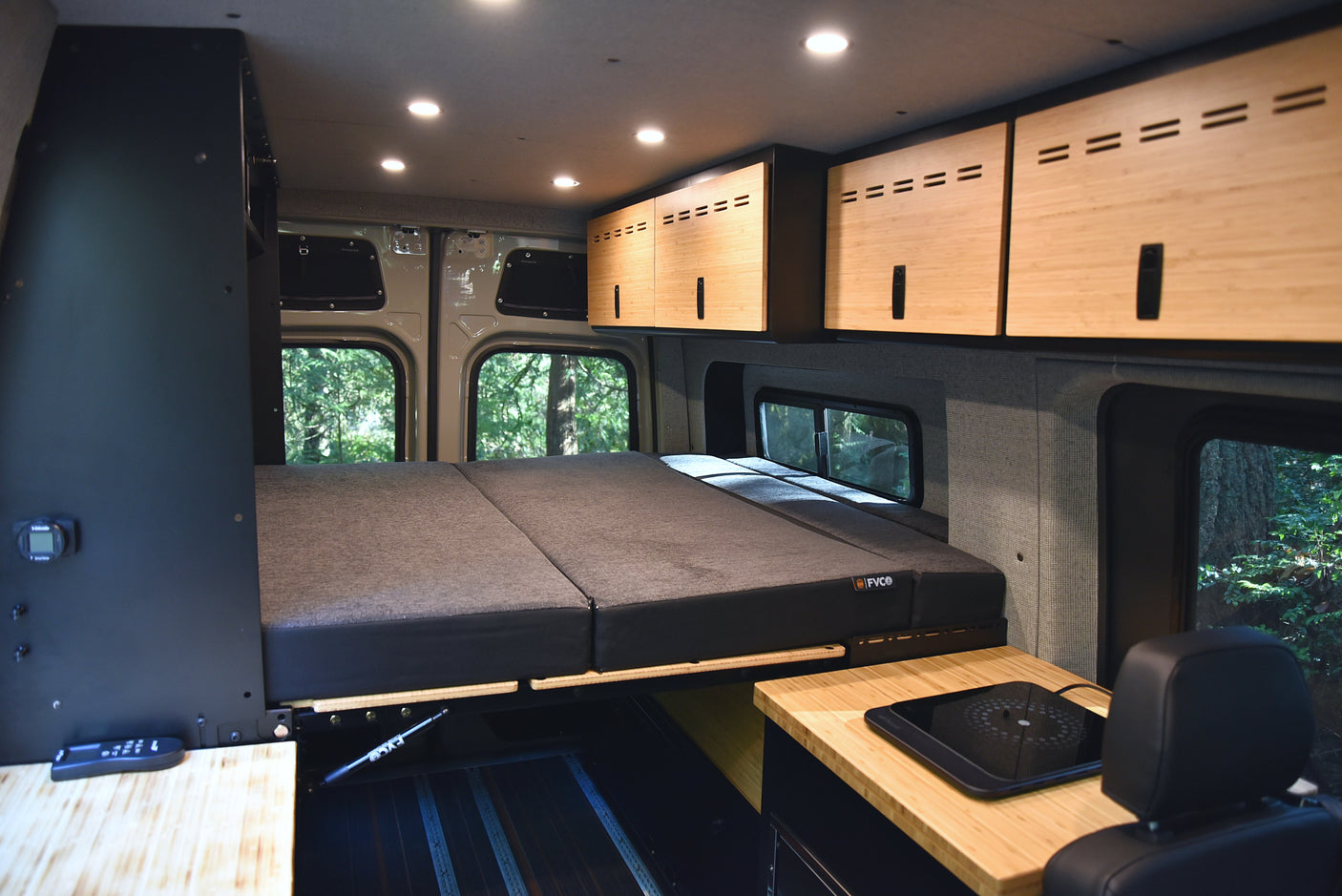 Interior of an RV rental camper van: comfortable bed, bamboo overhead storage, and kitchen area, with lush trees visible through the windows.