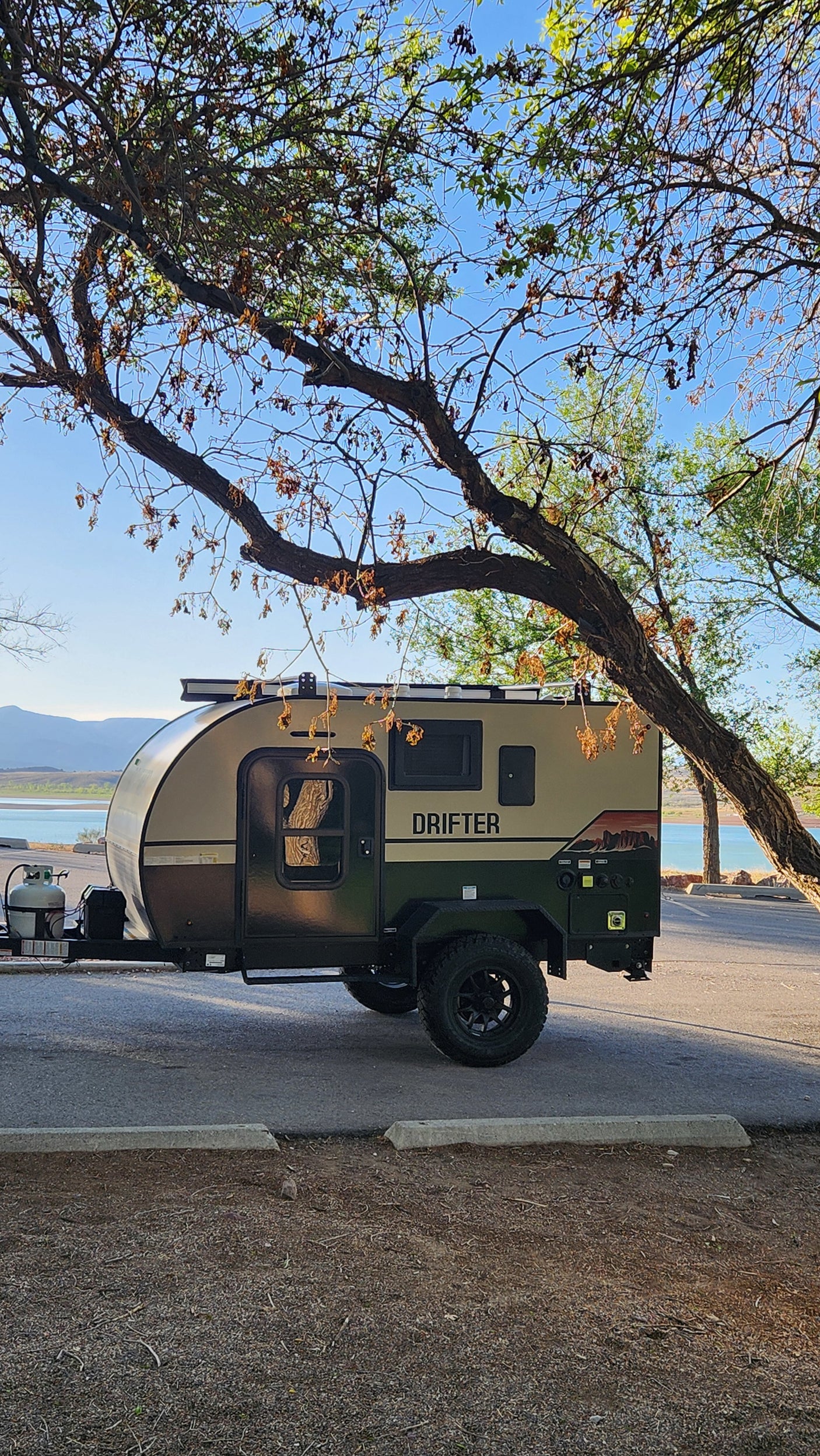 This Drifter RV rental camper is perfectly parked lakeside, under trees with mountains, ready for your next outdoor adventure.