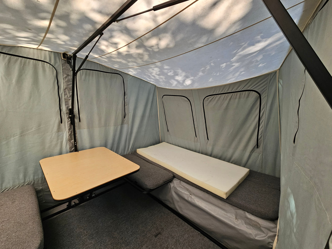 Inside an RV rental camper: light grey canvas interior with a wooden table, grey cushions, and a foam mattress.