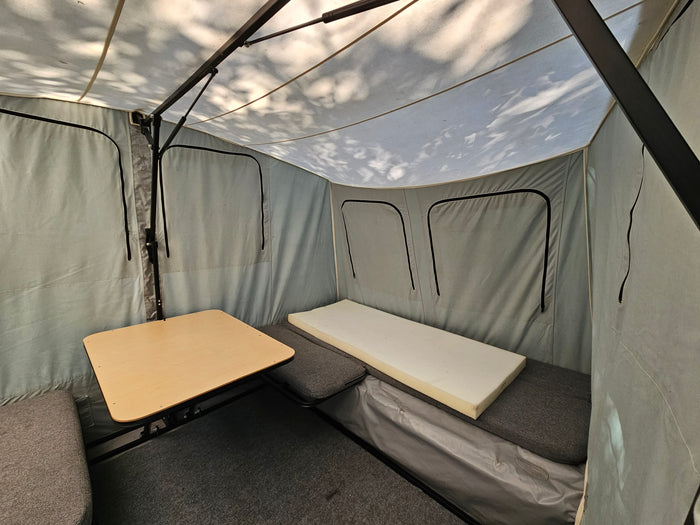 Inside an RV rental camper: light grey canvas interior with a wooden table, grey cushions, and a foam mattress.