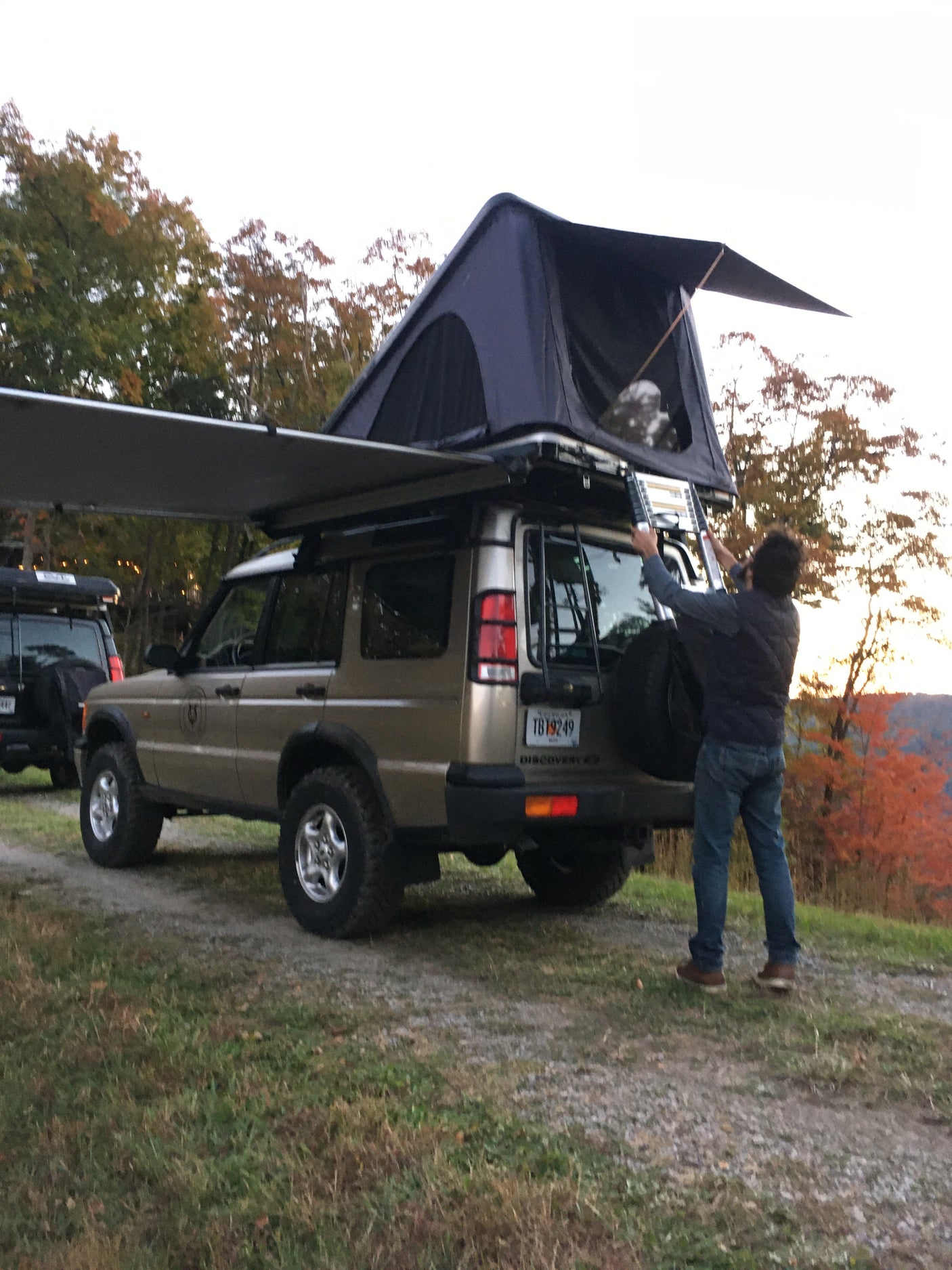 Adventure-ready RV rental: A gold Land Rover Discovery with a roof-top tent and awning deployed. A person sets up the ladder for camping.