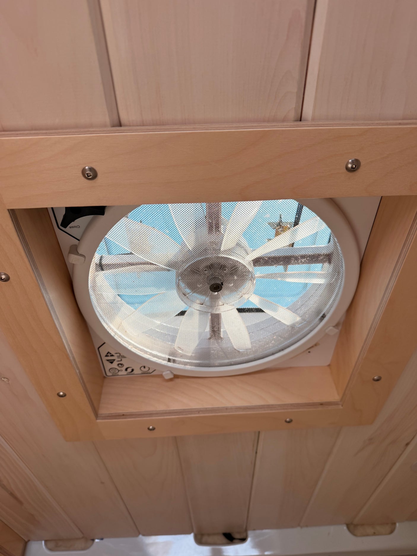 Inside an RV rental, a ceiling fan/vent with a mesh screen is framed by light wood panels, showing the sky above.