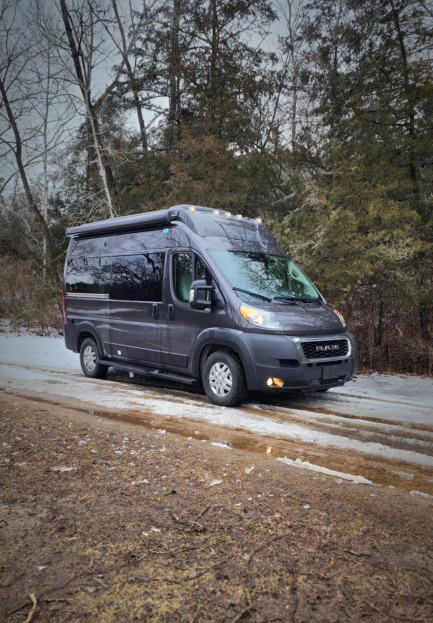 Dark grey camper van RV rental on a snowy, muddy path. Bare and evergreen trees line the winter road.