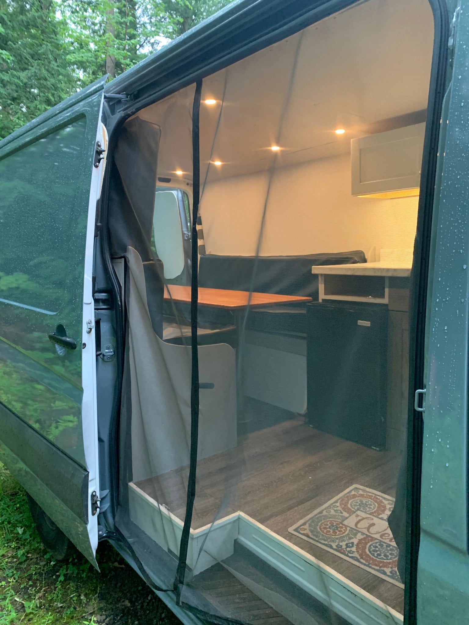RV rental with open side door and mosquito net, revealing a cozy camper van interior: bed, table, fridge, & "Welcome" mat.