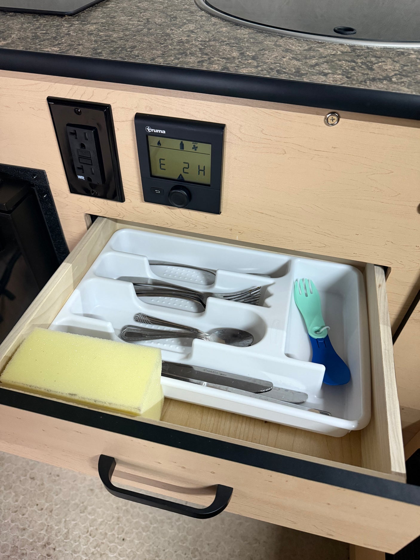 Open RV rental kitchen drawer: a cutlery organizer, sponge, & child's spork. A Truma control panel (E 2 H) & outlet are visible.