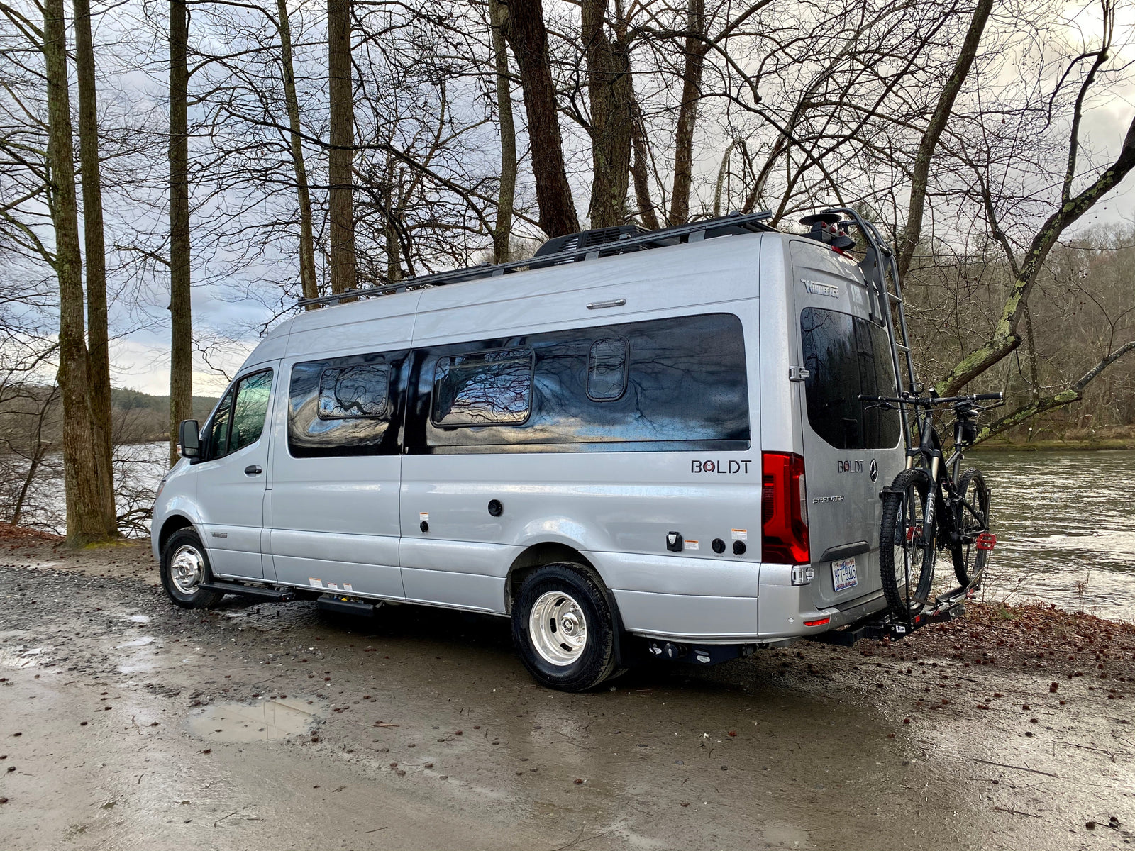 Silver Winnebago Boldt rv rental parked by a muddy river, ready for adventure with bikes on the back.