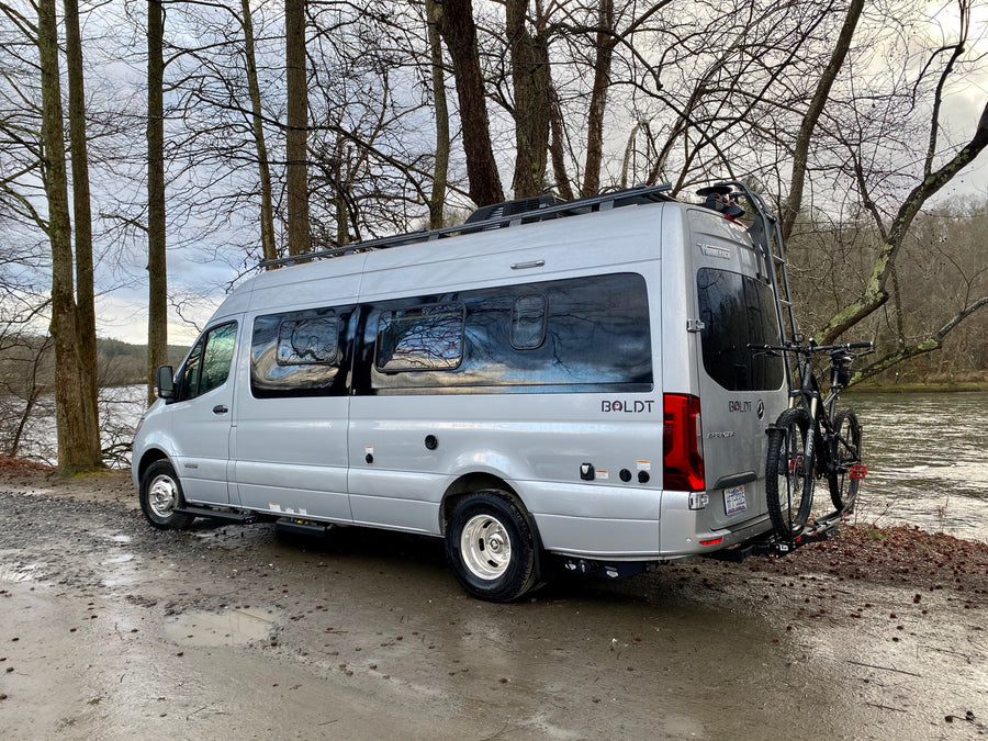 Silver Winnebago Boldt rv rental parked by a muddy river, ready for adventure with bikes on the back.
