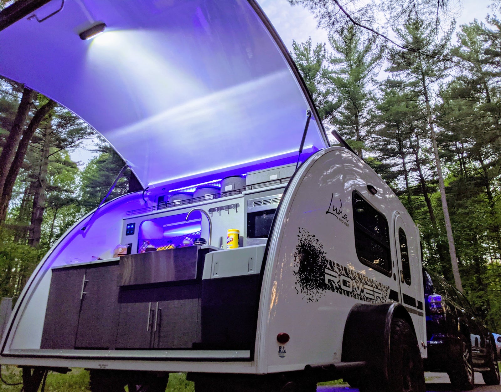 RV rental: A Luna Rover teardrop trailer's outdoor kitchen, lit with vibrant purple LEDs, open for camping in pine woods.