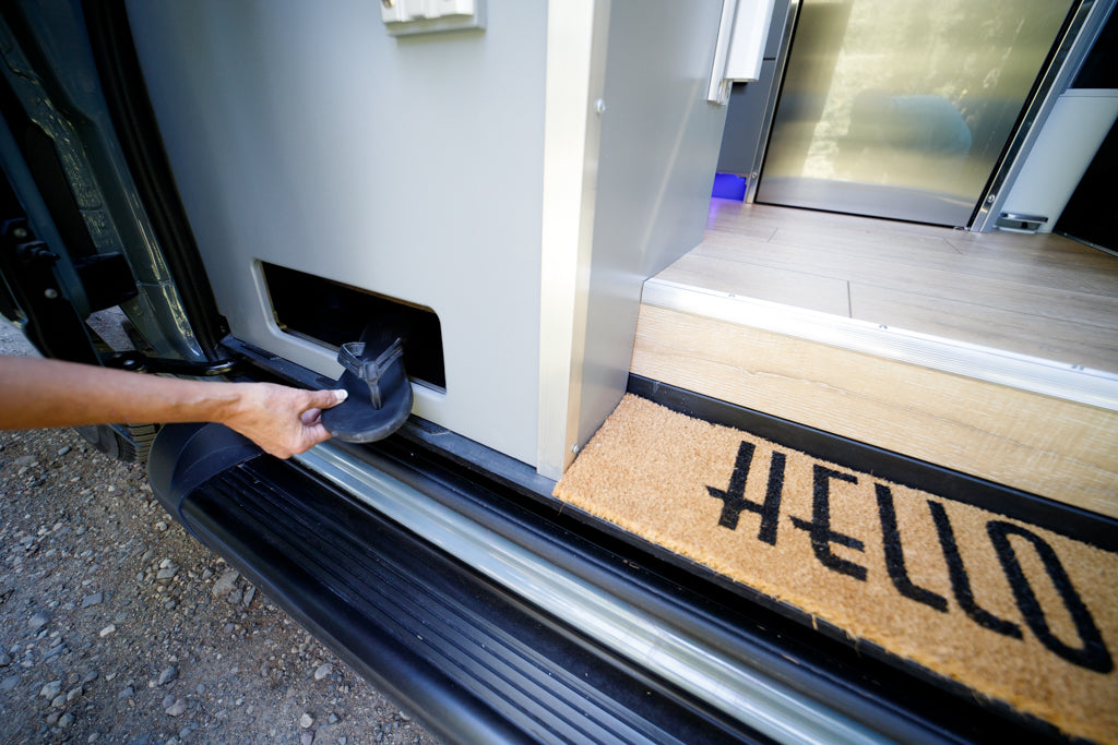 Organized RV rental entry: hand places flip-flop in built-in shoe storage slot by the "HELLO" welcome mat.