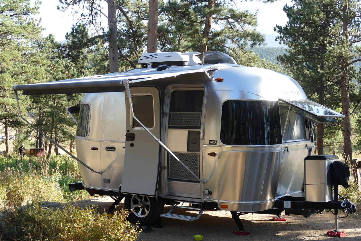 A shiny silver Airstream travel trailer, like an RV rental, is parked among tall pine trees and green brush, ready for outdoor adventure.