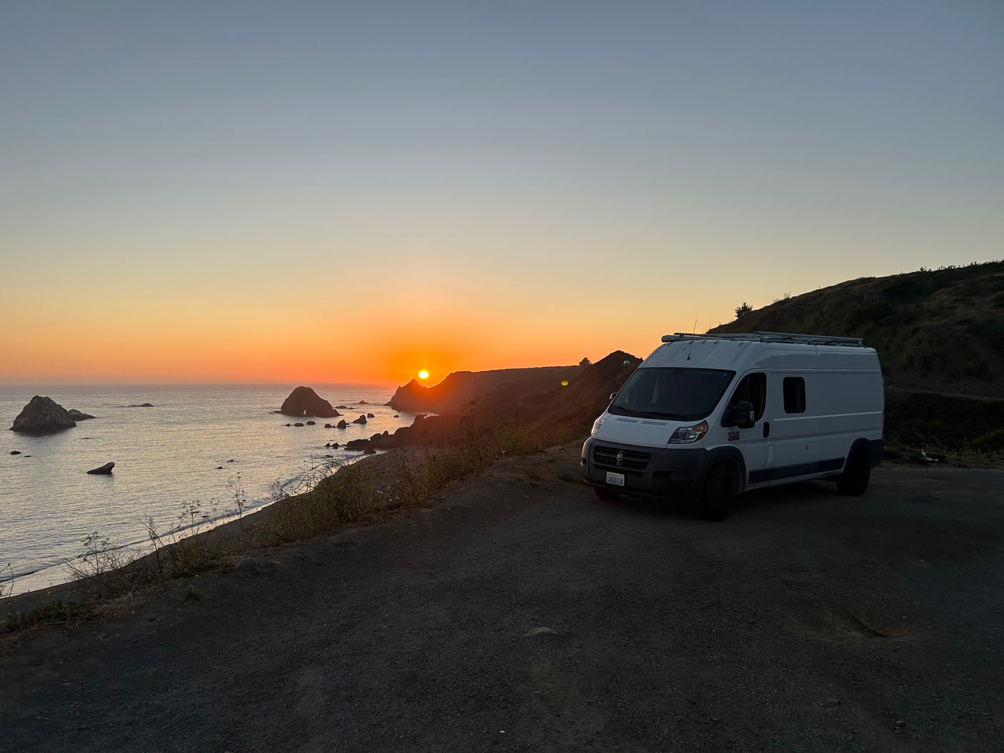 White RV rental parked at sunset by the ocean, with a vibrant sky and sea stacks. Ideal for a coastal road trip.