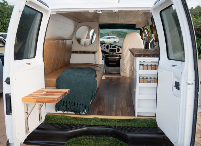 Inside this modern **rv rental** van: a cozy bench/bed, kitchen with spice rack, wood floors, and a fold-out table.