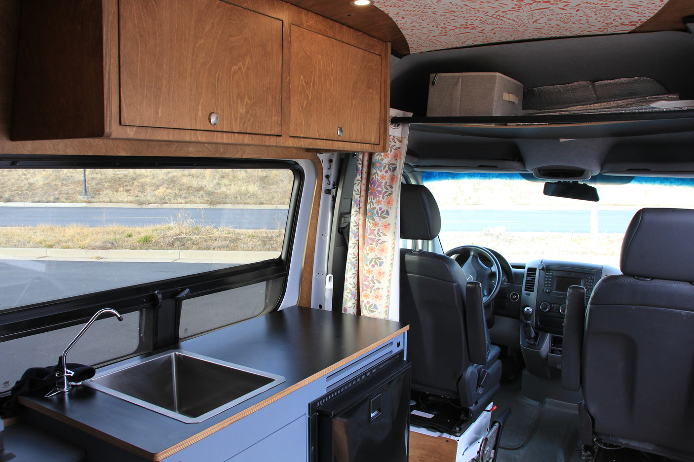 This RV rental features a camper van interior with a kitchenette, wooden overhead cabinets, and a view into the driver's cabin.