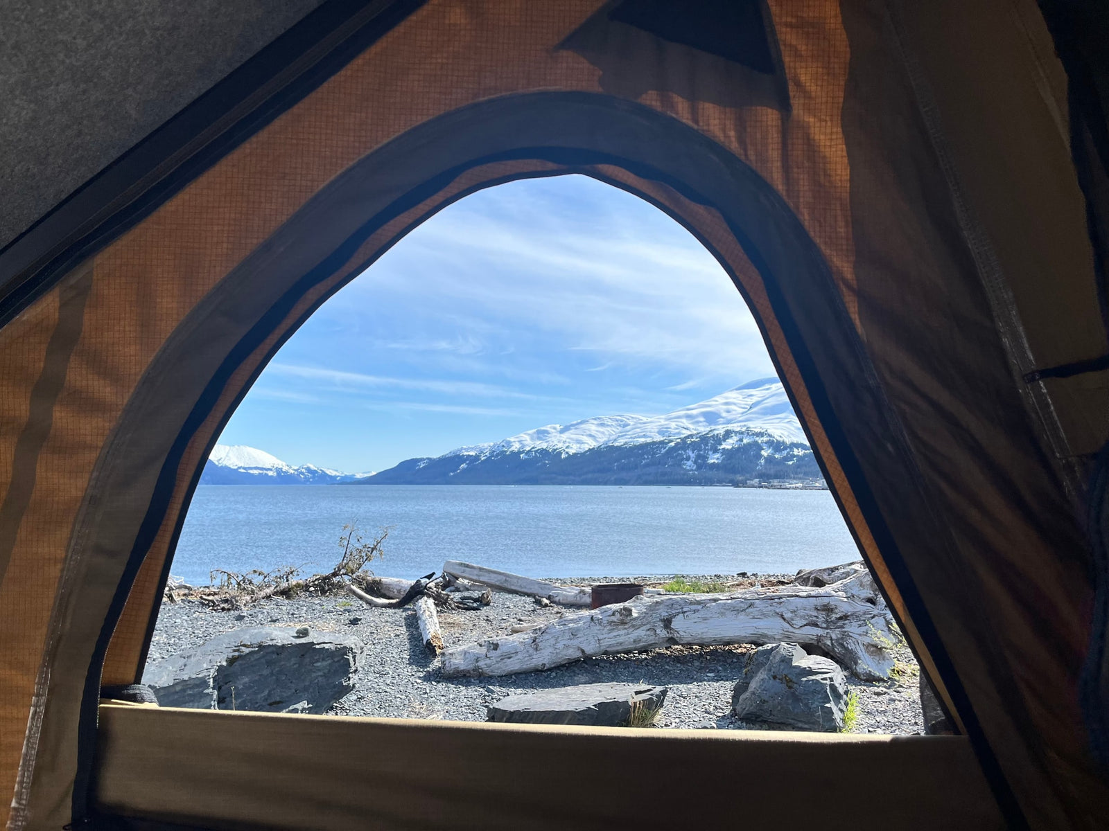 Serene view from inside an RV rental. A tranquil bay, snow-capped mountains, and a driftwood-strewn beach are visible through the opening.