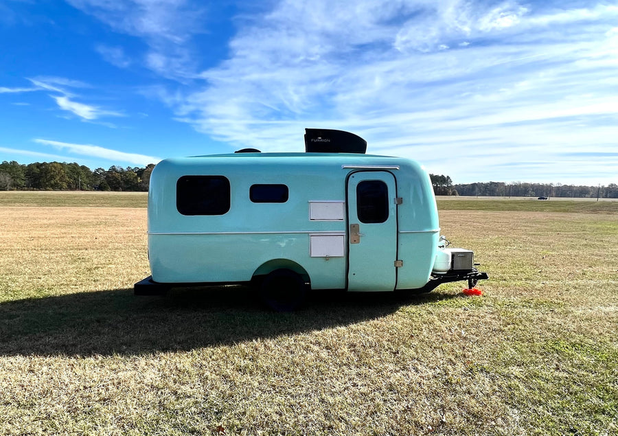 Light blue vintage-style travel trailer (rv rental) with a Furrion unit, parked in a sunny field under blue sky.
