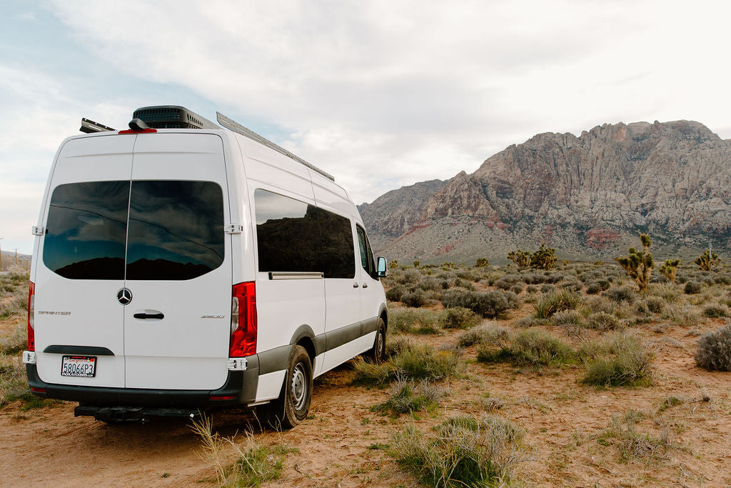 A white Mercedes Sprinter camper van RV rental parked in a vast desert landscape with rugged mountains under a cloudy sky.