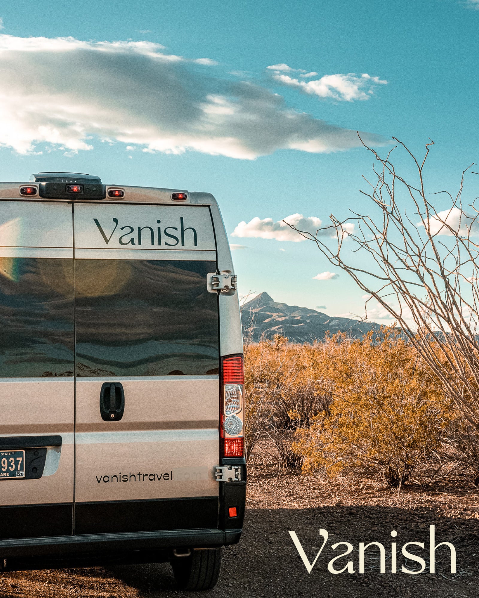 Rear view of a silver Vanish RV rental campervan parked in a desert landscape with mountains and blue sky.