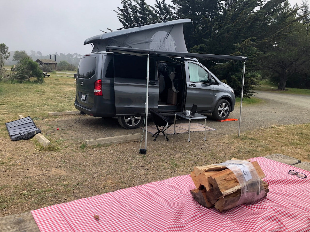 Enjoying an RV rental adventure! Grey pop-top camper van set up at a scenic campsite with solar power & firewood.