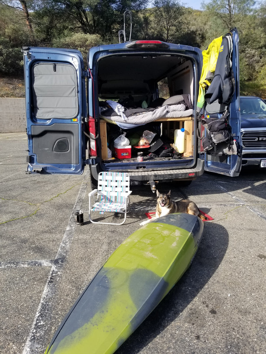 Blue rv rental van prepped for adventure: bed, gear, kayak, and a smiling dog. Ready for a great outdoor escape.