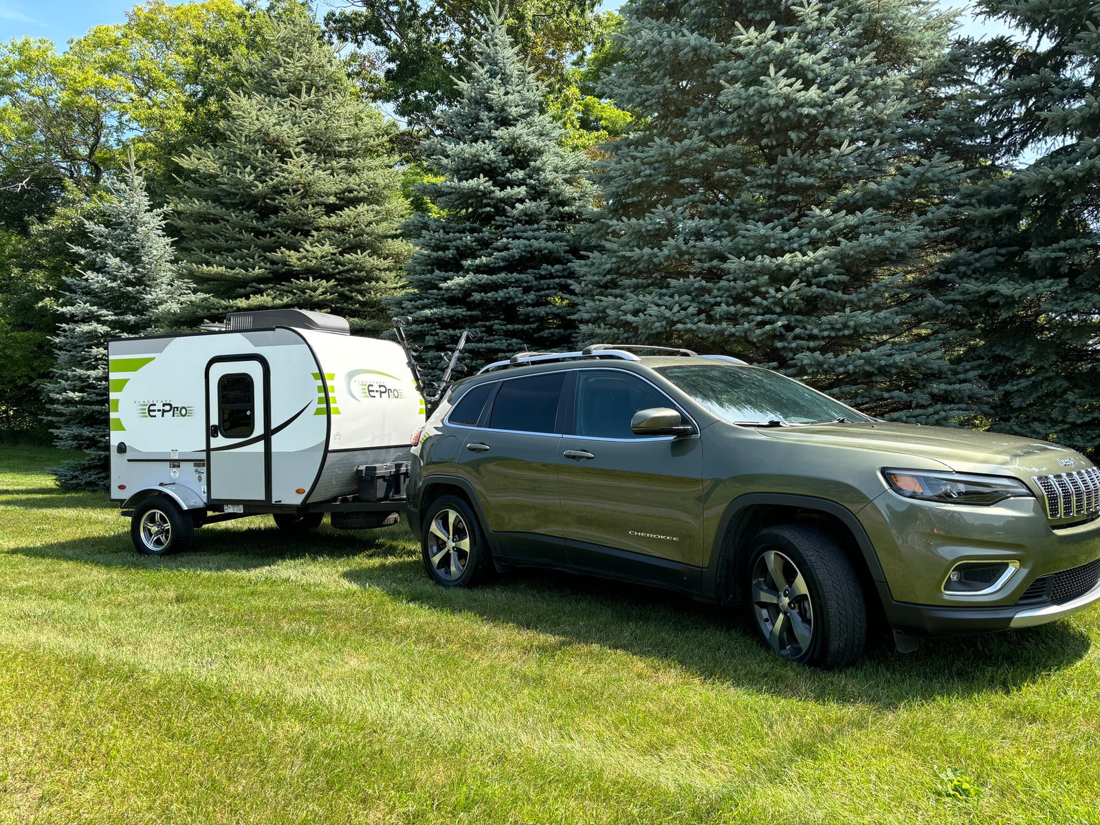 This RV rental features a green Jeep Cherokee SUV towing a white Flagstaff E-Pro travel trailer on sunny grass near trees.