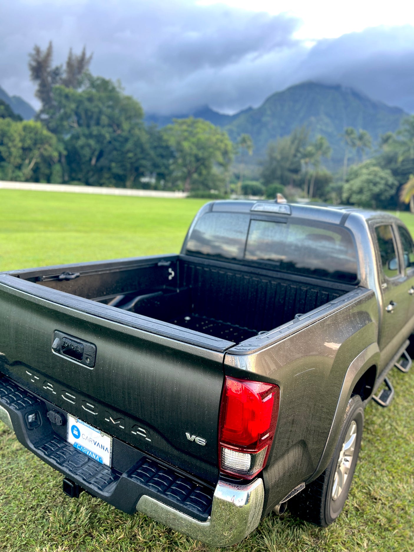 A dark gray Toyota Tacoma pickup truck rests on vibrant green grass, with lush mountains shrouded in clouds behind. Ready for exploration or an RV rental adventure.