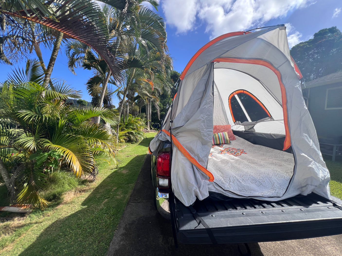 This truck bed tent transforms a pickup into a cozy, portable RV rental for camping. Lush palm trees line the sunny drive.