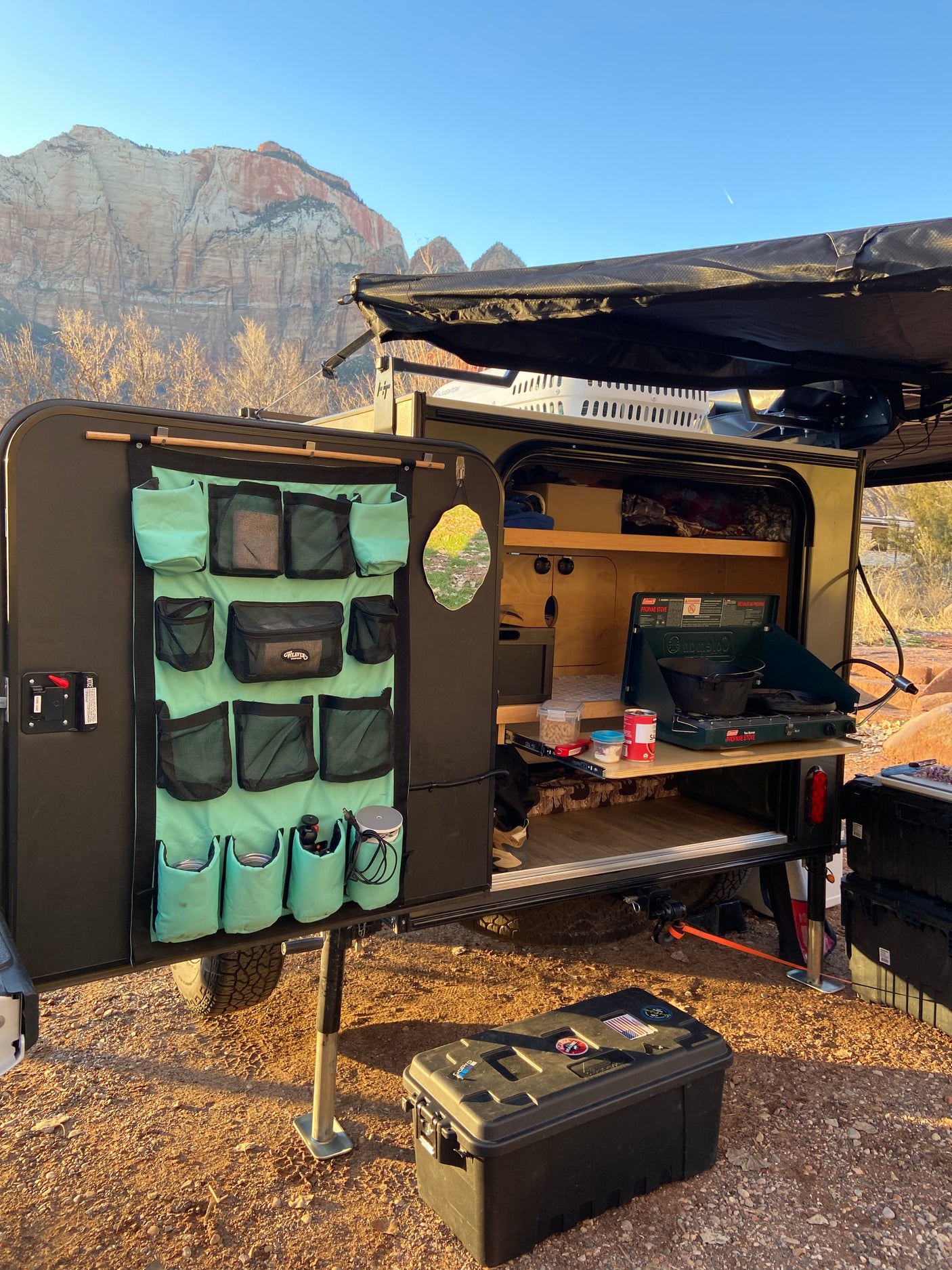 An RV rental camper's open outdoor kitchen with a stove and organizer, set against majestic mountains at sunrise.