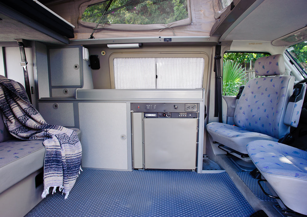 RV rental interior: pop-top camper with a compact kitchen, built-in fridge, patterned seats, and a cozy blanket.