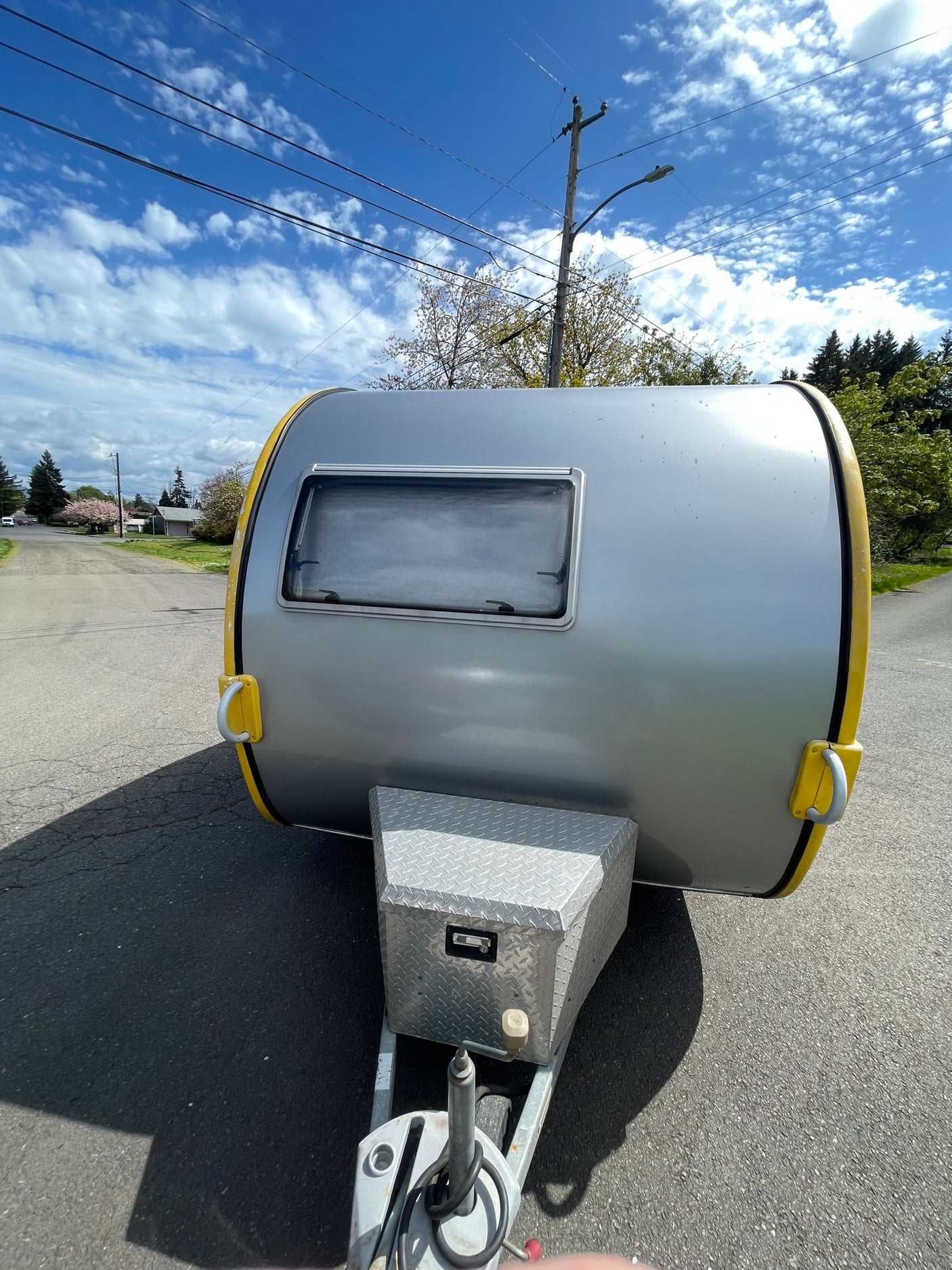 Front view of a sleek silver teardrop RV rental, featuring yellow trim and a storage box, parked on an asphalt road.