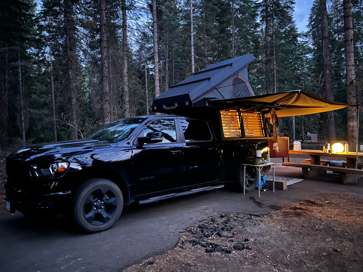 Black truck with Alu-Cab camper & rooftop tent at a lit forest campsite. Ideal for overlanding or an RV rental adventure.