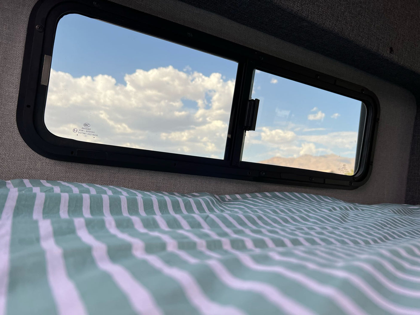 RV rental bed offers a view of blue sky, white clouds, and distant mountains through its twin windows.