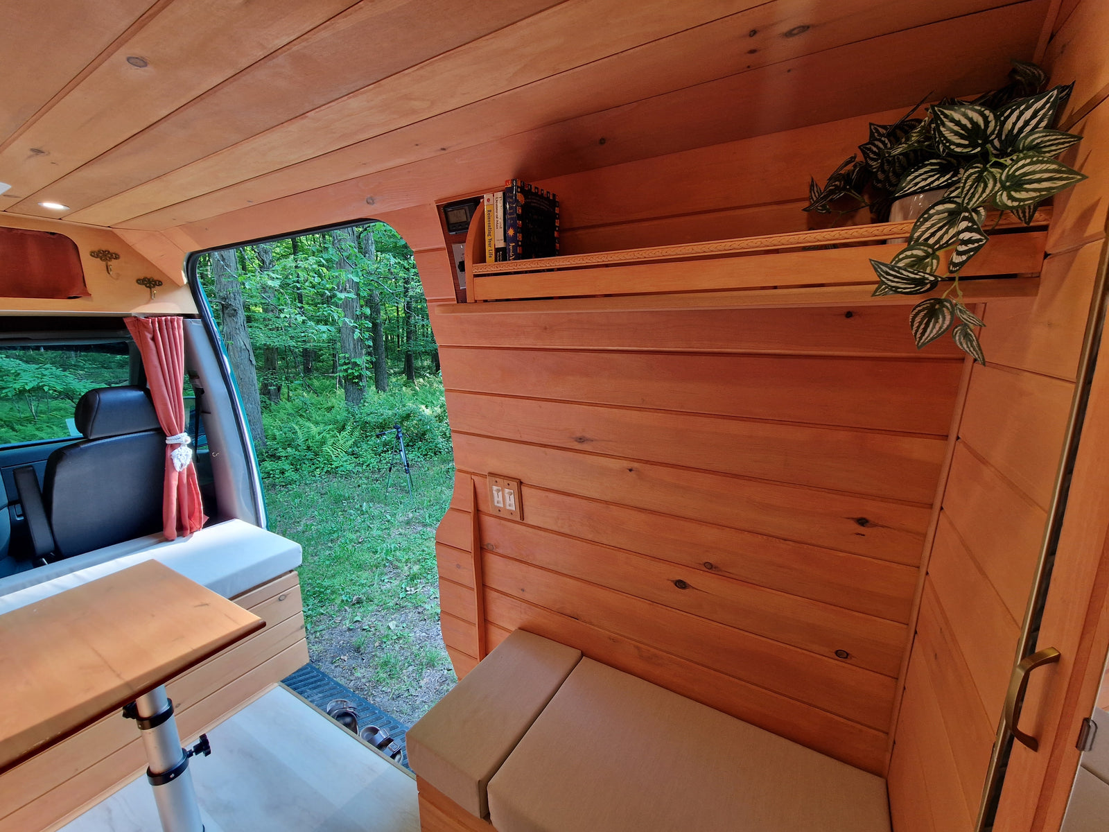 Wood-paneled RV rental interior, showing cozy seating, a bookshelf with plants, and a forest view from the open door.